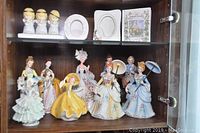 Porcelain figurines displayed on a shelf in a cabinet, showing various women in period dresses and hats with parasols.