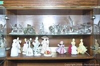 Glass and porcelain figurines arranged in cabinet display with glass animal figurines on top shelf and female figurines with Avon bottles and bells on lower shelf.