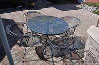 Photo of round metal mesh outdoor table with clear glass top and four matching metal mesh chairs on a brick patio in daylight.