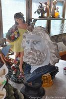 Closer view of the male bust figurine in shades of white and blue, female figurine with bouquet, and other figurines on table under daylight.