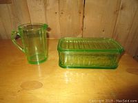 Two uranium glass items displayed on a wooden table: one measuring cup with handle and spout, one rectangular covered dish.