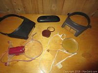 Various magnifying glasses including two headwear magnifiers, handheld magnifier with red frame, compact magnifier in brown case, and small clear handheld magnifier with string, placed on wooden surface.