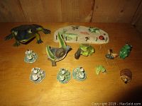 Photo showing a variety of small frog decorations and figurines including a large frog, several small frogs in different poses, a frog on a leaf, three frogs on lily pads, and a hedgehog figurine.