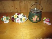 Four items on wooden table: three porcelain flower arrangements and one ceramic biscuit jar with floral design