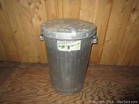 Front view of the metal garbage can showing its cylindrical shape, side handles, and attached identification sticker.