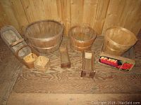 Photo showing a variety of wooden bushel baskets and assorted small wooden boxes and crates arranged on wooden floor with plywood wall background.