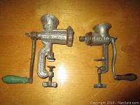 Two vintage manual meat grinders side by side on a wooden surface showing metal bodies and wooden handles, one labeled Dominion and one labeled Wizard.