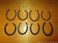 All eight horseshoes arranged on a wooden surface, showing four black-painted and four rusty horseshoes.