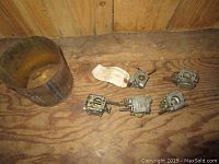 Five small carburetors arranged on a wood surface with a metal bucket nearby.