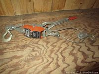 Come along tool placed on wooden surface showing hooked ends, lever handle with orange grip, and cable system.