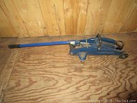Blue hydraulic car jack with long handle and wheels on wooden floor, showing side view