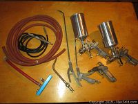 Overview of lot showing two paint spray guns with metal cups, two coiled hoses, two spray lance extensions, and one air blow gun on a wooden table.