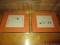 Two framed vintage prints by Fischer displayed on wooden surface, showing colorful illustrations on beige paper with orange mats.