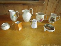 Eight vintage porcelain pitchers and creamers with distinct floral and scenic designs displayed on wooden surface. Includes a variety of shapes and decorative styles indicating vintage porcelain collectors' items.