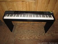 Full view of Casio Privia digital keyboard in black with stand, placed on wooden floor against wood paneling.