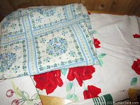 Photo showing a folded tablecloth with blue and green floral block pattern over a white tablecloth with large red roses.