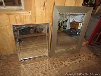 Two mirrors leaning against a plywood wall: larger one with matte grey wooden frame, smaller one with reflective mirrored frame.