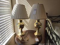 Two matching brass table lamps on a wooden side table near window blinds, highlighting lampshades and reflective brass bases.