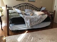 Full view of daybed with wooden posts and black metal decorative headboard and side rail, showing linens, pillows and bottom pull-out bed.