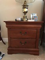 Front view of wooden nightstand showing two drawers, metal handles, and decorative molding on top edges. Medium brown finish.