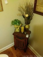 Wicker cabinet with faux greenery in angel pot, gold vase with dried branches, pillar candle, and small glass tealight holder on top