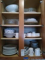 Cabinet showing shelves of assorted dinnerware including bowls, plates, cups and mugs in various patterns and plain white.