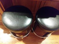 Top-down view of two round black cushioned bar stools with wooden legs and metal footrests on a wood floor.