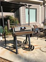 Front side view of large metal smoker grill with wheels and firebox under a patio cover