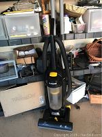 Eureka vacuum cleaner shown standing on floor with shelves and boxes behind it.