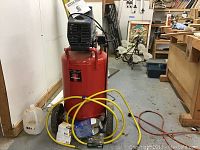 Full view of red vertical tank compressor on wheels with attached yellow air hose and power cord
