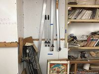 Five aluminum bar bench clamps hanging vertically on a pegboard wall. Clamps appear used and vary in length. Part of a woodworking bench is visible nearby.