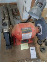 Front facing photo of orange 8 inch bench grinder with dust on surface, lamp attached, grinding wheels on either side, and several tool rests on the table beside it.
