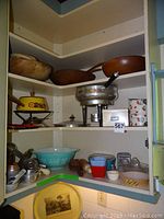 Full view of corner shelf with wooden salad bowls on top shelf, yellow enamel fondue set with burner and stand, Pyrex bowl, metal food mill, measuring cups, graters, and other small kitchen utensils