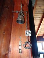 Wall-mounted brass bell from side angle showing mount, chain, and spoon beneath.