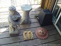 Overview photo of garden items on porch including cement statue, pedestal, plastic planters, sun face decoration and floral tile.