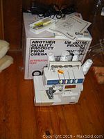 Front view of Omega 334D serger machine with spools of thread, instruction manual, accessories, and box behind.