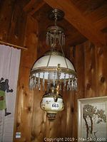 Full view of hanging oil lantern showing floral decorated white glass shade with hanging crystal prisms and brass hardware hanging from ceiling mount.