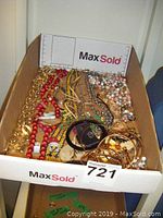 Full view of mixed costume jewelry items inside a cardboard box showing various chains, bead necklaces, and bracelets mainly in gold and red tones.