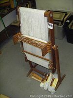 Front view of wooden Guildcraft Thackery Junior B knitting loom with white yarn and partially finished knit product. Extra yarn skeins and wooden knitting tools placed at base.