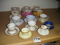 Full display of 9 assorted teacups with matching saucers and 5 demitasse cups with saucers arranged on cardboard surface.