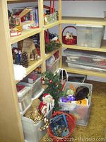 Shelves and floor area with multiple plastic bins containing assorted Christmas decorations including ornaments, small trees, baskets, and figurines.