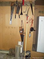 Hand tools including various hand saws, pruning clippers, pole saw, light, some wood, all hanging and leaning against a wall in a workshop area.