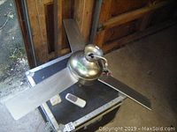 Ceiling fan laying on a case with visible three metallic blades and remote control beside it in a dimly lit room.