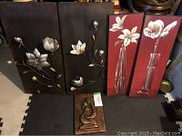 Five flower-themed wall art pieces displayed on a black mat, including two large dark metal panels with silver flowers, two burgundy floral paintings, and one small bronze piece.