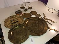 Full view of the lot items including three large brass plates with detailed engravings, two metal bird sculptures, a vintage balance scale with two hanging bowls, and some small brass weights.