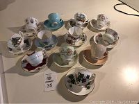 Overview of all eleven assorted teacups with matching saucers arranged on flat surface showing varied floral and scenic designs.
