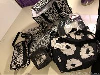 Overview of assorted Thirty One bags with black and white floral and damask patterns, including tote bags and a small pouch with a camera design.