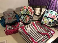 Multiple colorful Thirty One bags and Roots backpack on table, showing patterns and styles