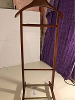 View of the entire wooden valet stand with its hanger and frame against a white and purple fabric background.