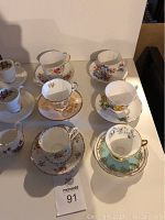 View showing all seven teacups and saucers arranged on table with various floral and gold designs.